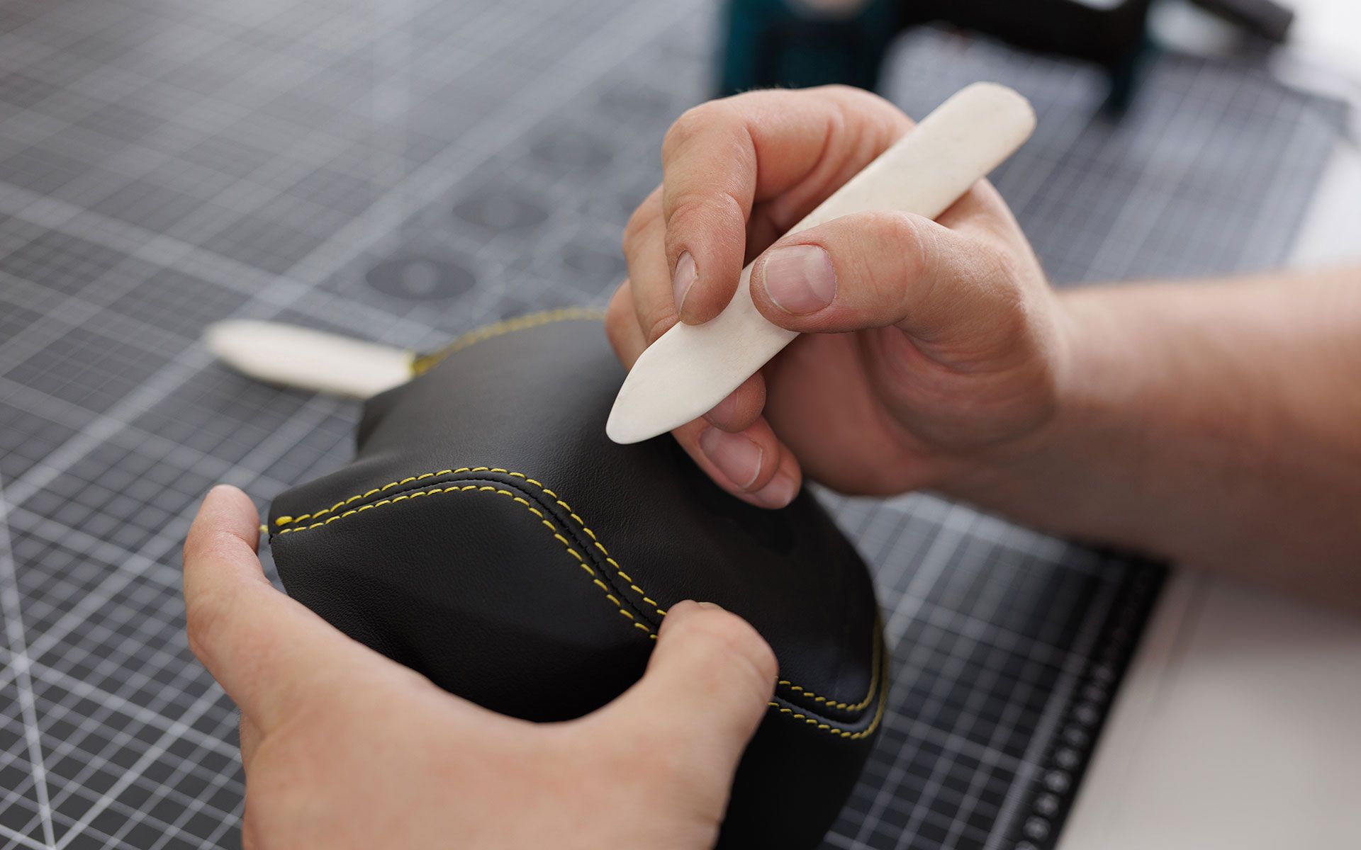 Hands using a bone folder to smooth leather with yellow stitching. Crafting on a grid mat.