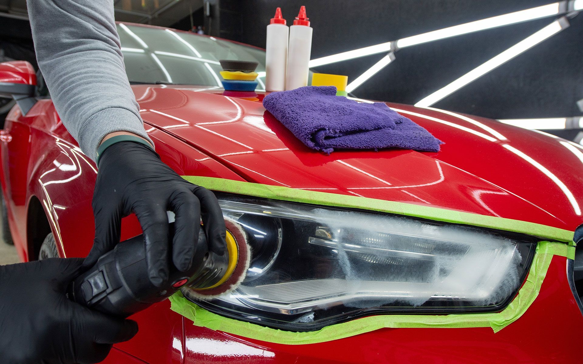 Person polishing car headlight, red car in auto detailing shop.