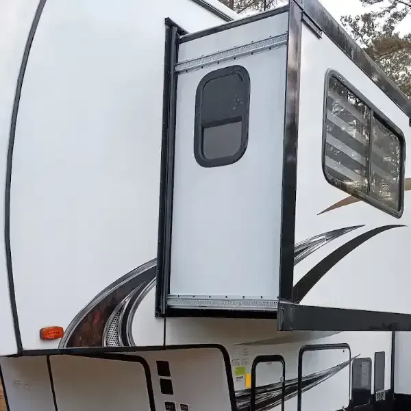 White RV with slide-out extended, featuring a small window and a larger window with horizontal blinds.