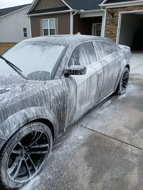 Gray car covered in white foam being washed in a driveway.