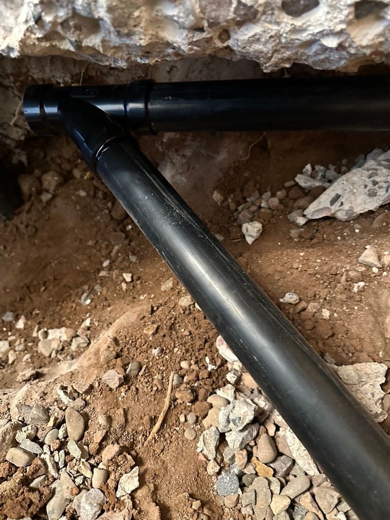 Black pipes in a trench under a concrete structure; dirt and gravel surround the pipes.