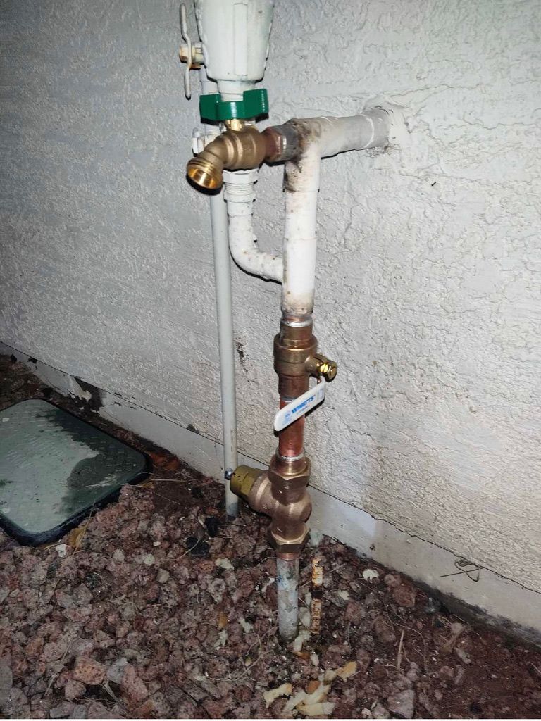 Outdoor water pipes and valves, including a green handled valve and a white meter. Brown dirt surrounds the pipes.