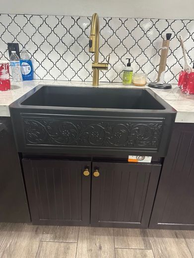 Black kitchen sink with ornate front, gold faucet, and dark cabinets.