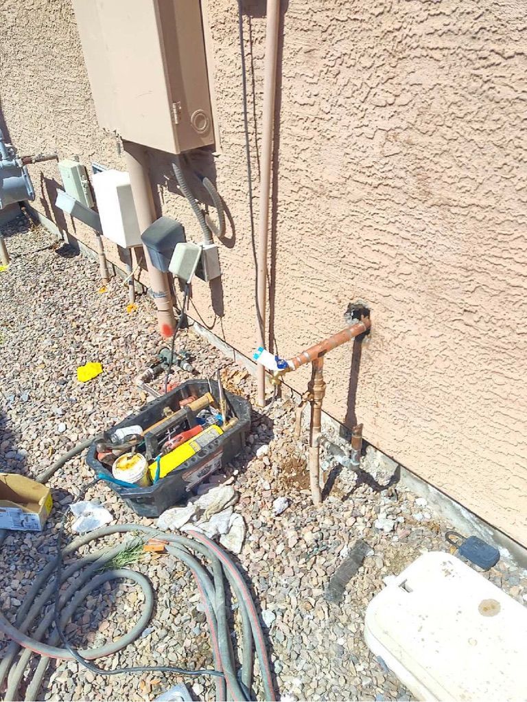 Plumbing tools and pipes next to a stucco wall, with a toolbox, and gravel ground.
