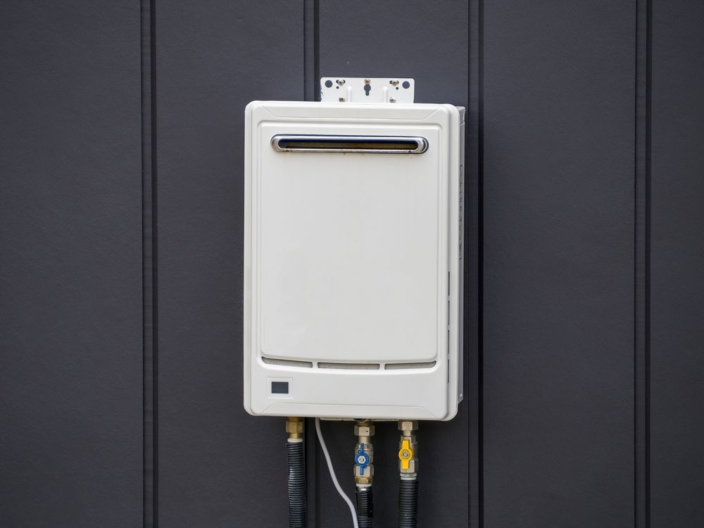 White tankless water heater mounted on a dark gray exterior wall with black and metal pipes.