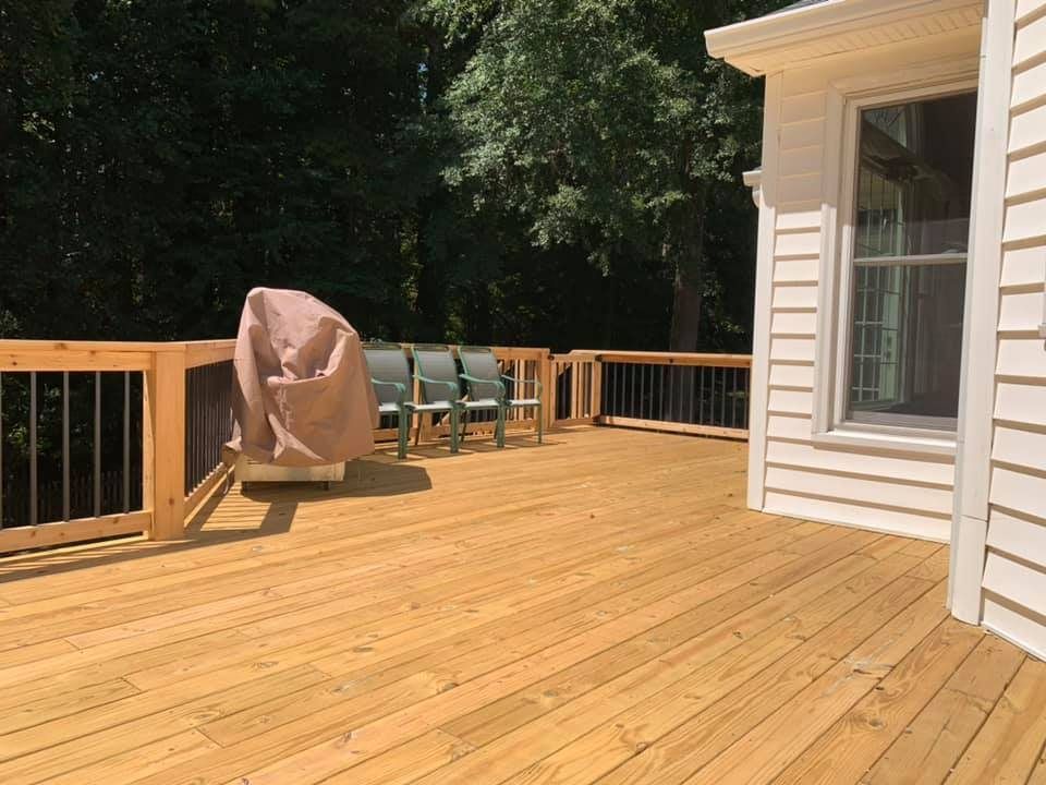 Wooden deck with chairs, grill, and railing next to a white house, surrounded by trees.