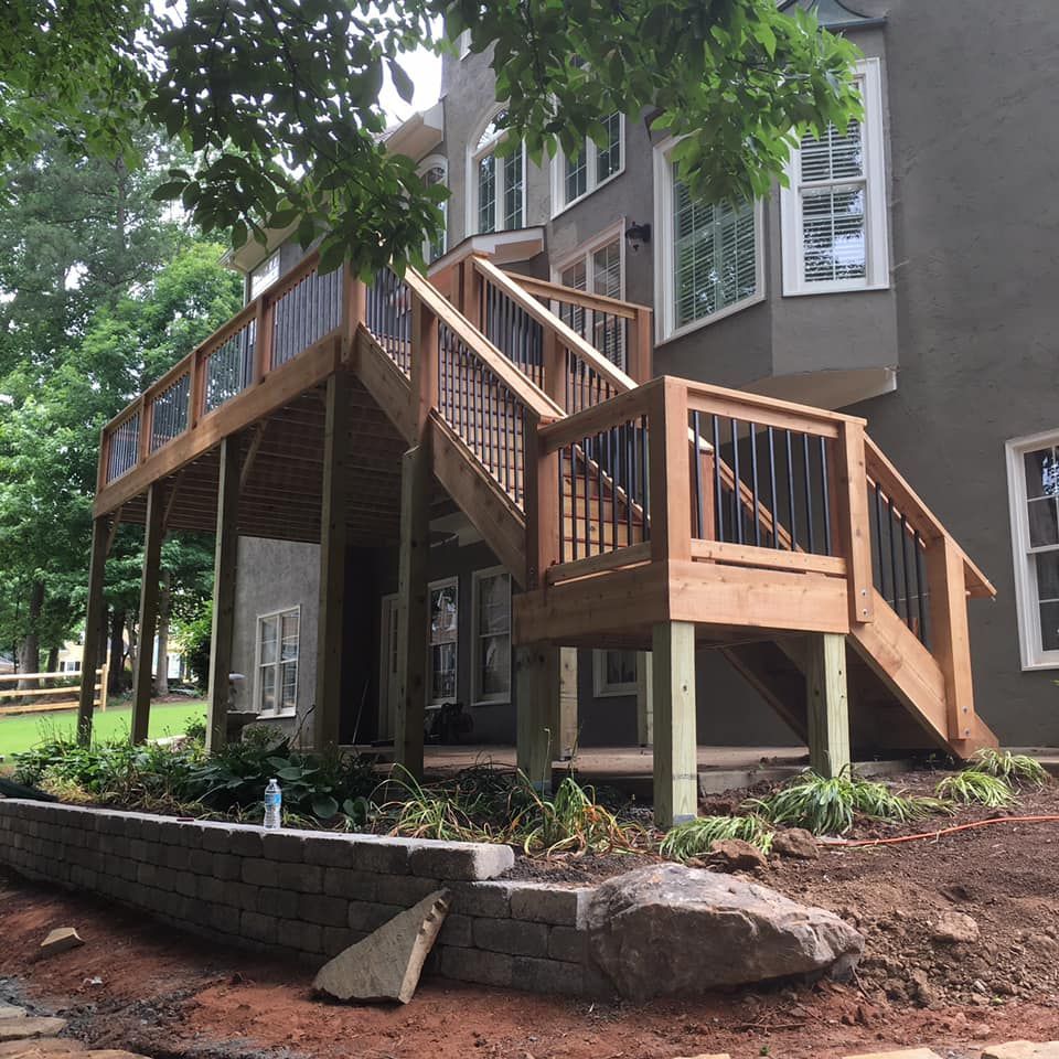 Wooden deck with stairs, attached to a gray house, with a stone retaining wall and landscaping in front.