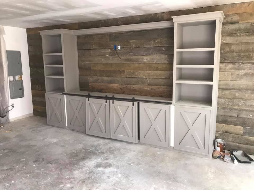Built-in gray cabinet unit with open shelving, barn door style lower cabinets, and a rustic wood-plank wall in a home.