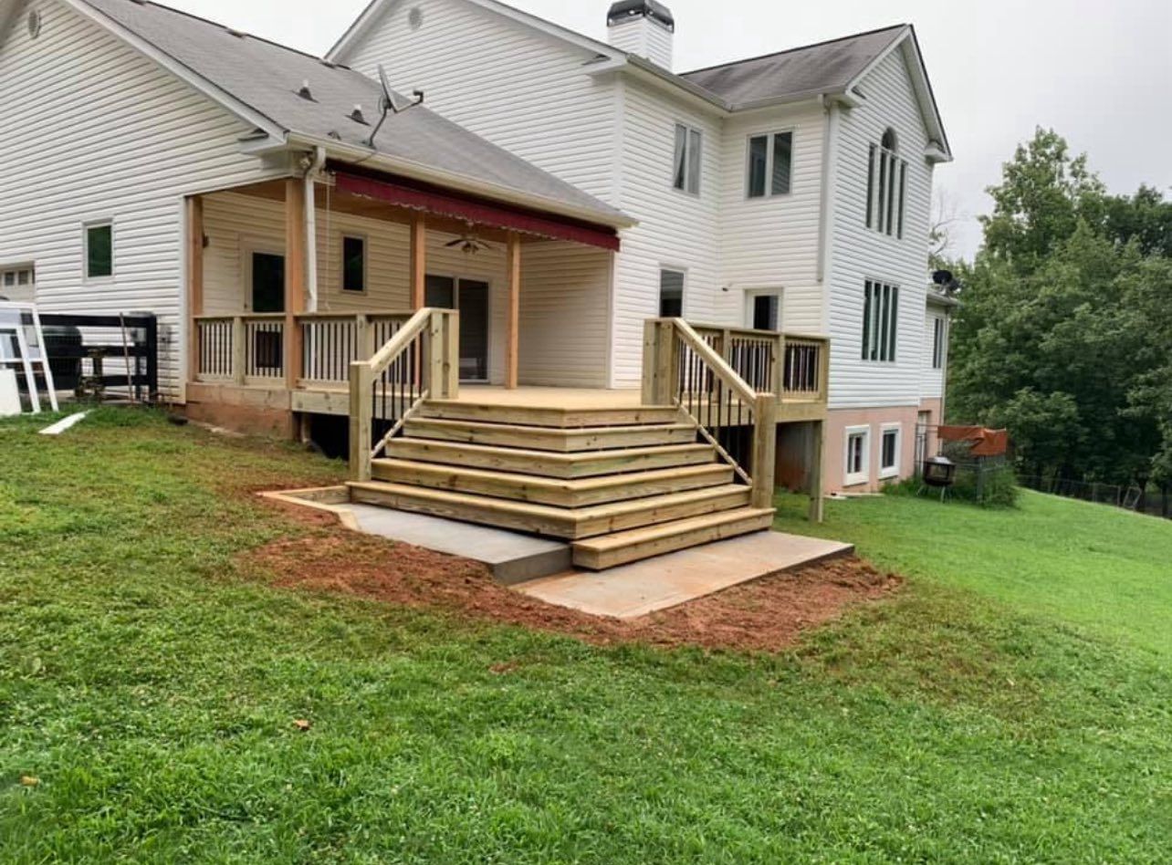 Wooden deck with steps leading to a concrete patio, attached to a white house on a grassy hill.