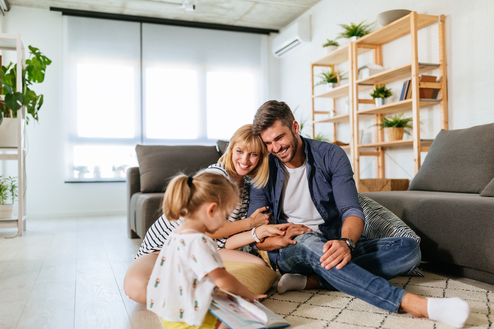 Une famille est assise sur un tapis dans un salon, souriante, en train de lire un livre ensemble dans une maison moderne et lumineuse.