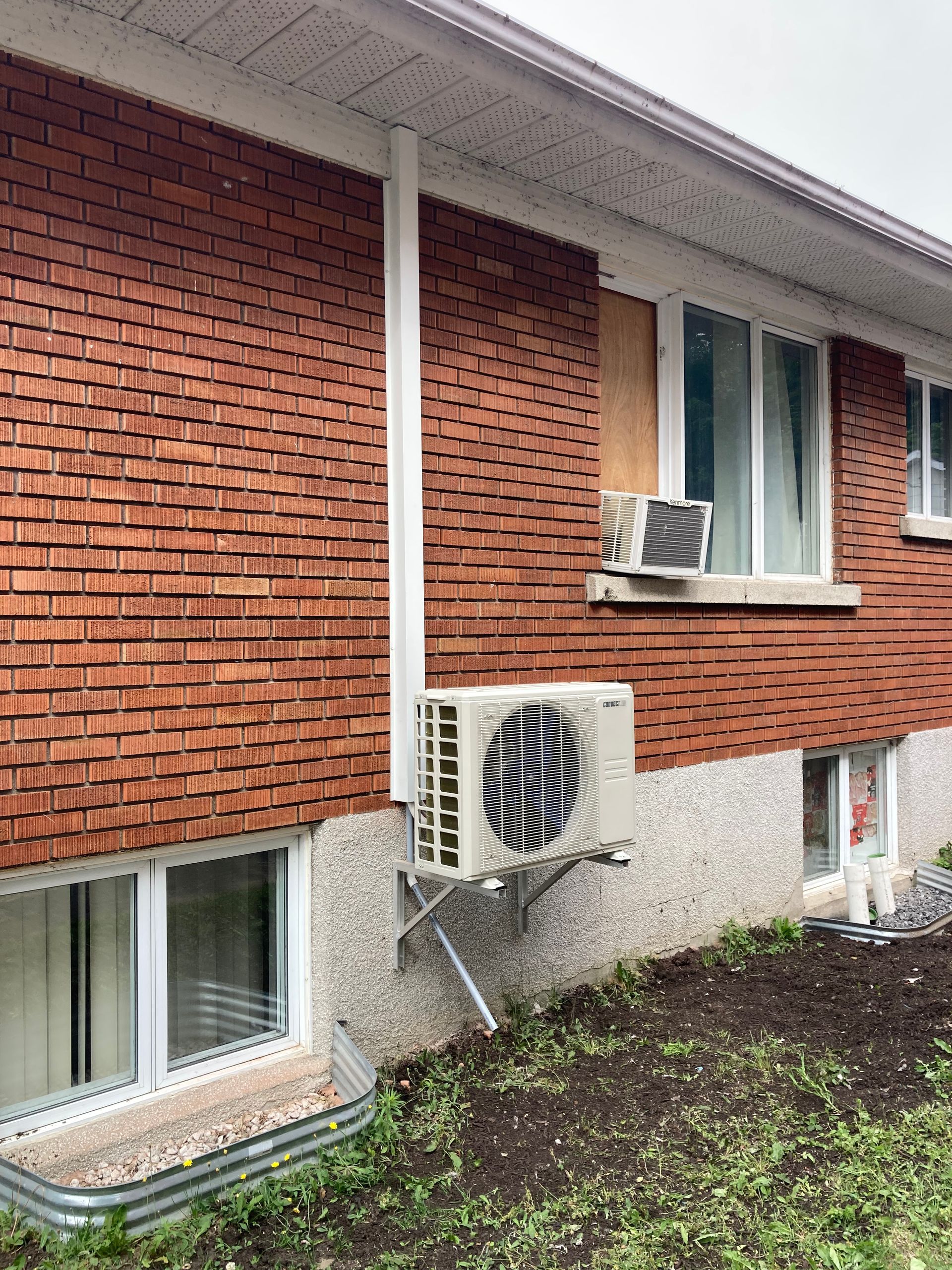 Un mur extérieur en briques rouges avec un climatiseur de fenêtre, une unité extérieure murale et une fenêtre au niveau inférieur.