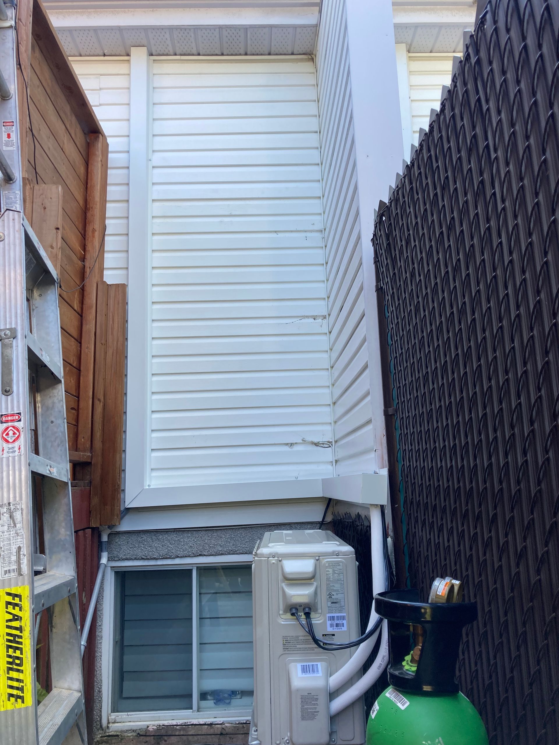 Vue extérieure étroite montrant un revêtement blanc, une fenêtre de sous-sol, une unité de chauffage, ventilation et climatisation, une échelle et un écran d'intimité sombre.