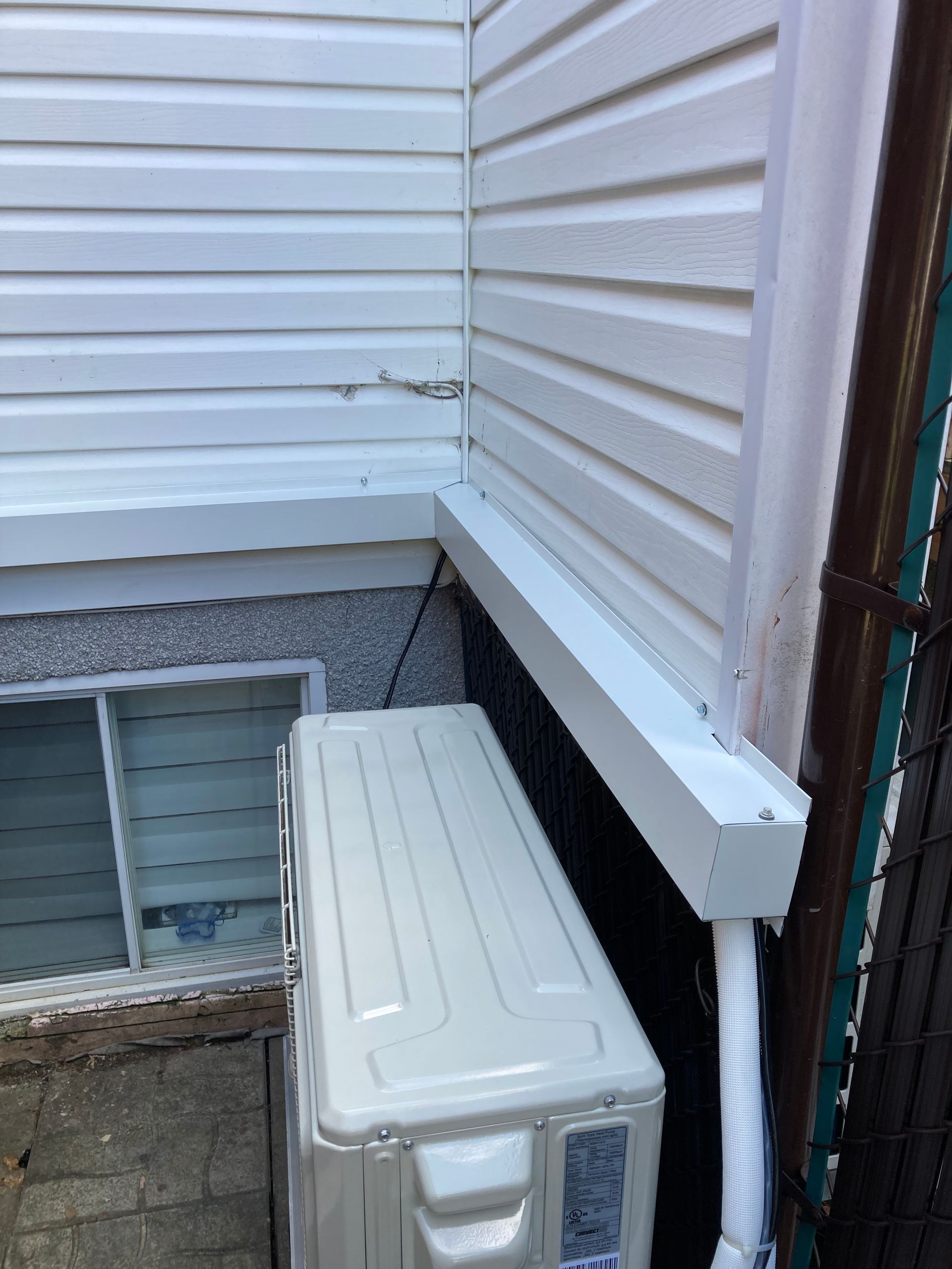 Un climatiseur extérieur est installé contre un mur de maison, avec un revêtement blanc et un tuyau recouvert courant le long de sa base.