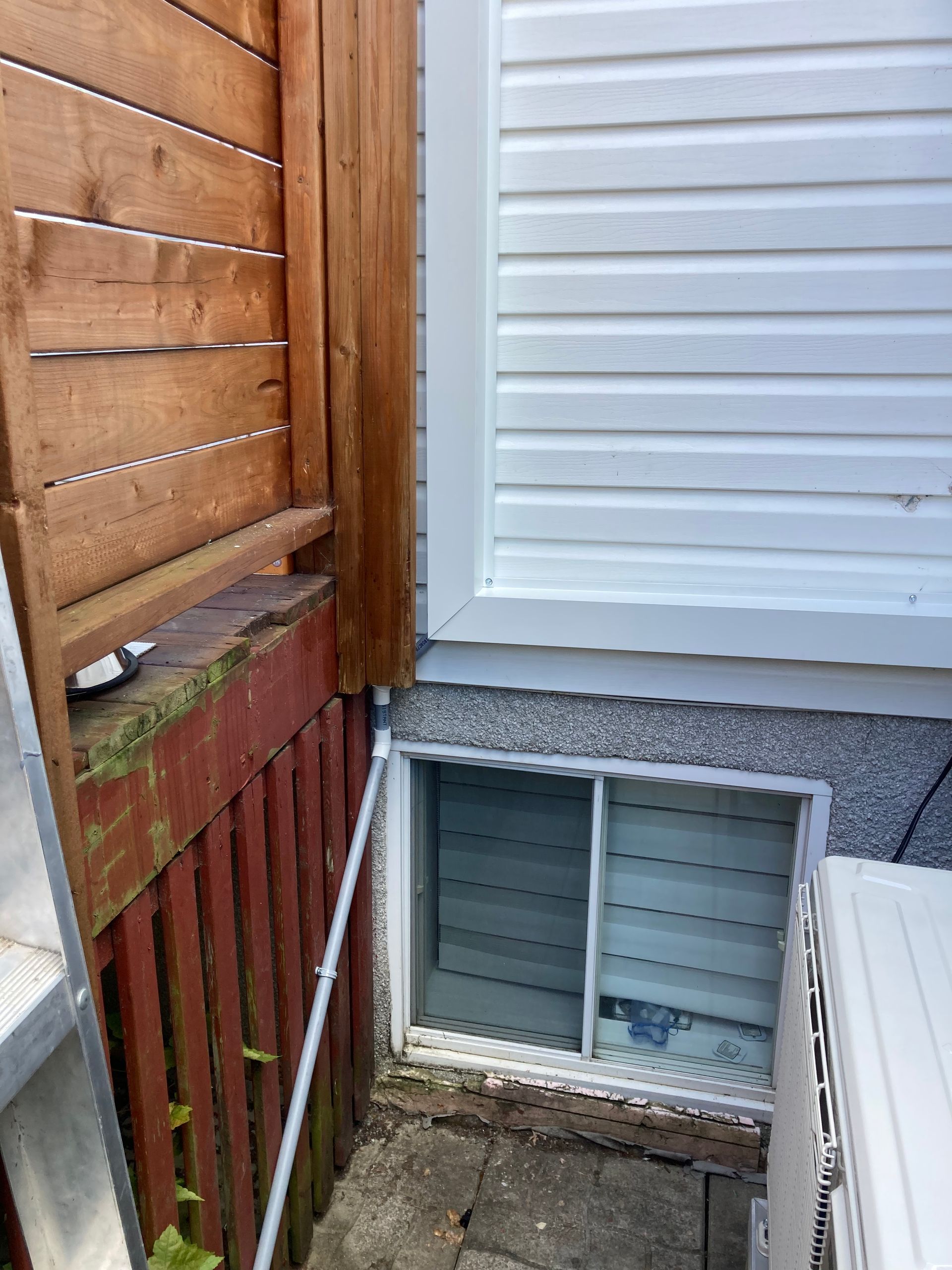 Vue au niveau du sol des fondations d'une maison montrant une fenêtre de sous-sol à persiennes à côté d'une clôture en bois et d'un mur.