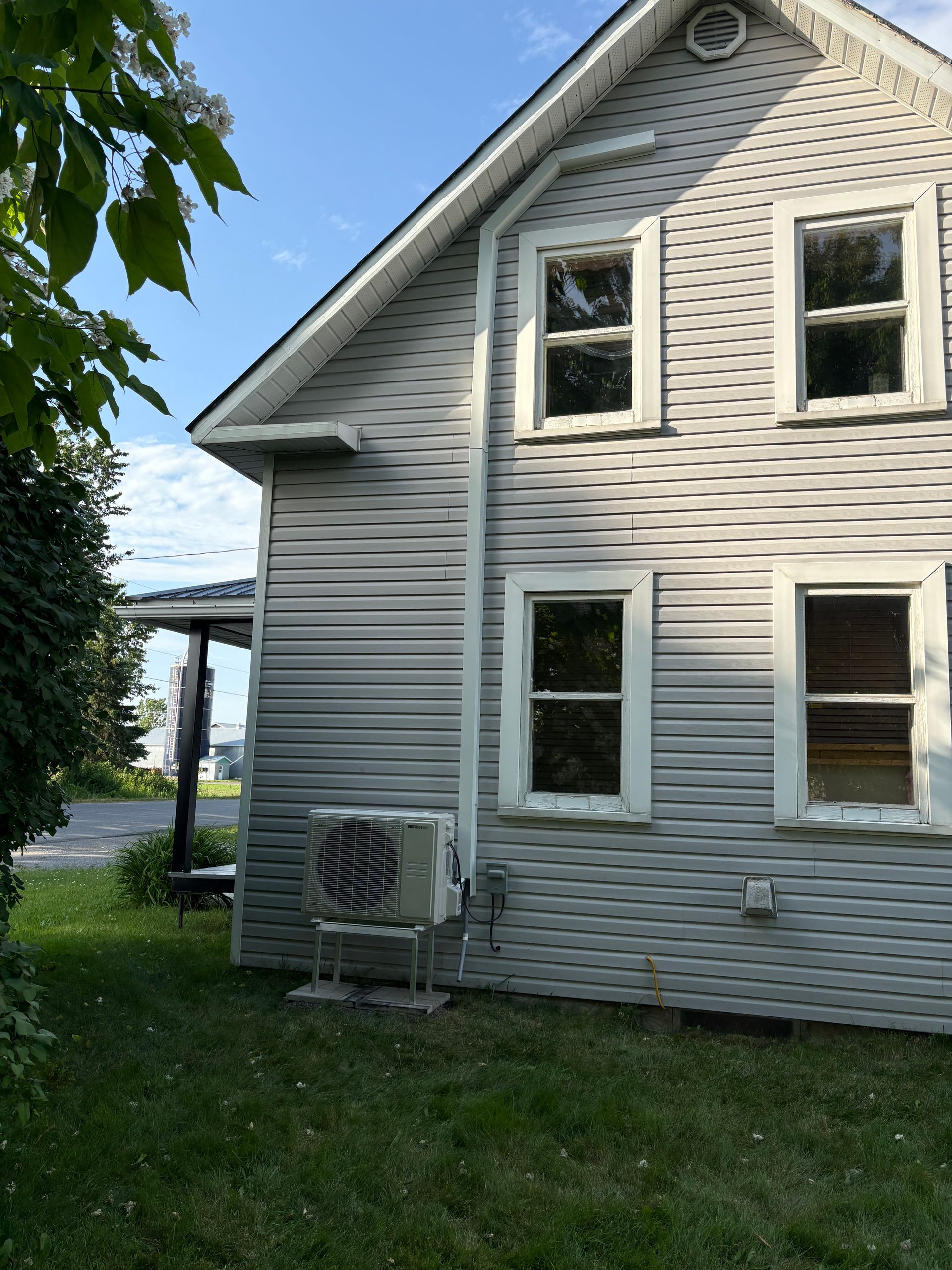 Vue extérieure latérale d'une maison claire à deux étages avec un bardage horizontal et un climatiseur extérieur sur la pelouse.