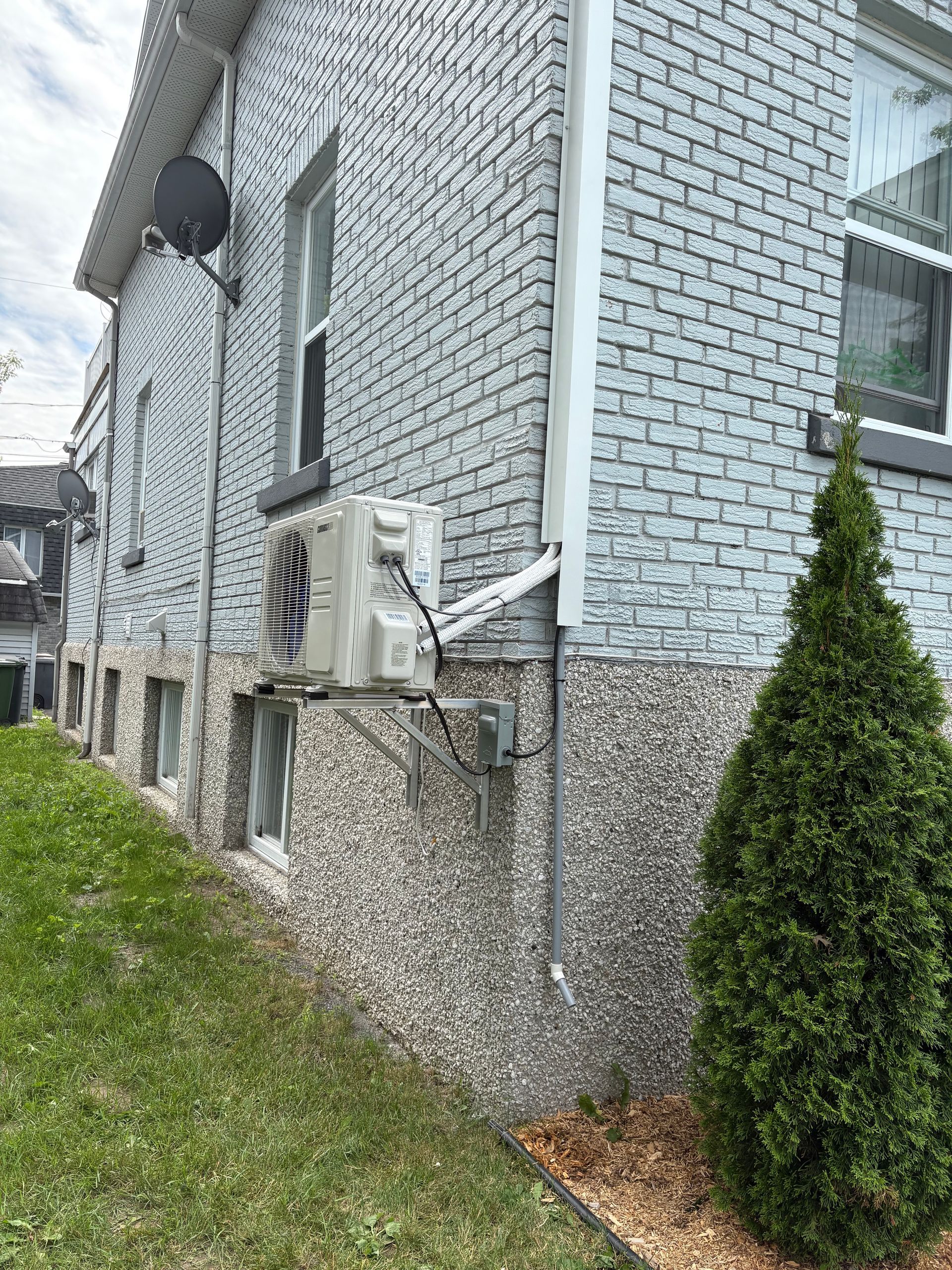 Vue latérale d'une maison aux murs de briques grises, aux fondations en galets, avec un climatiseur extérieur et un petit pin.