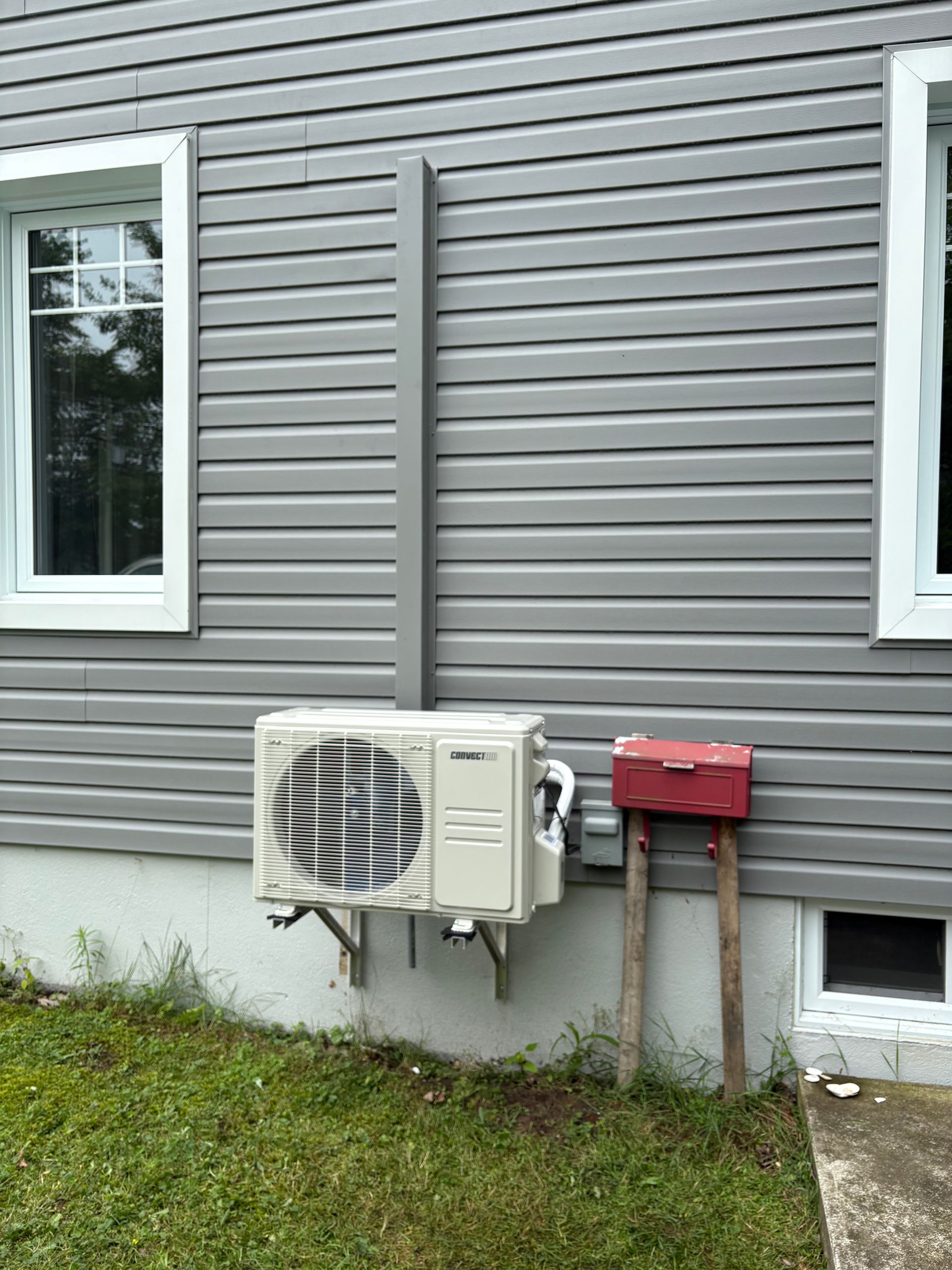 Un climatiseur split extérieur monté sur un mur bardé gris, à côté d'une boîte aux lettres rouge sur un poteau en bois.