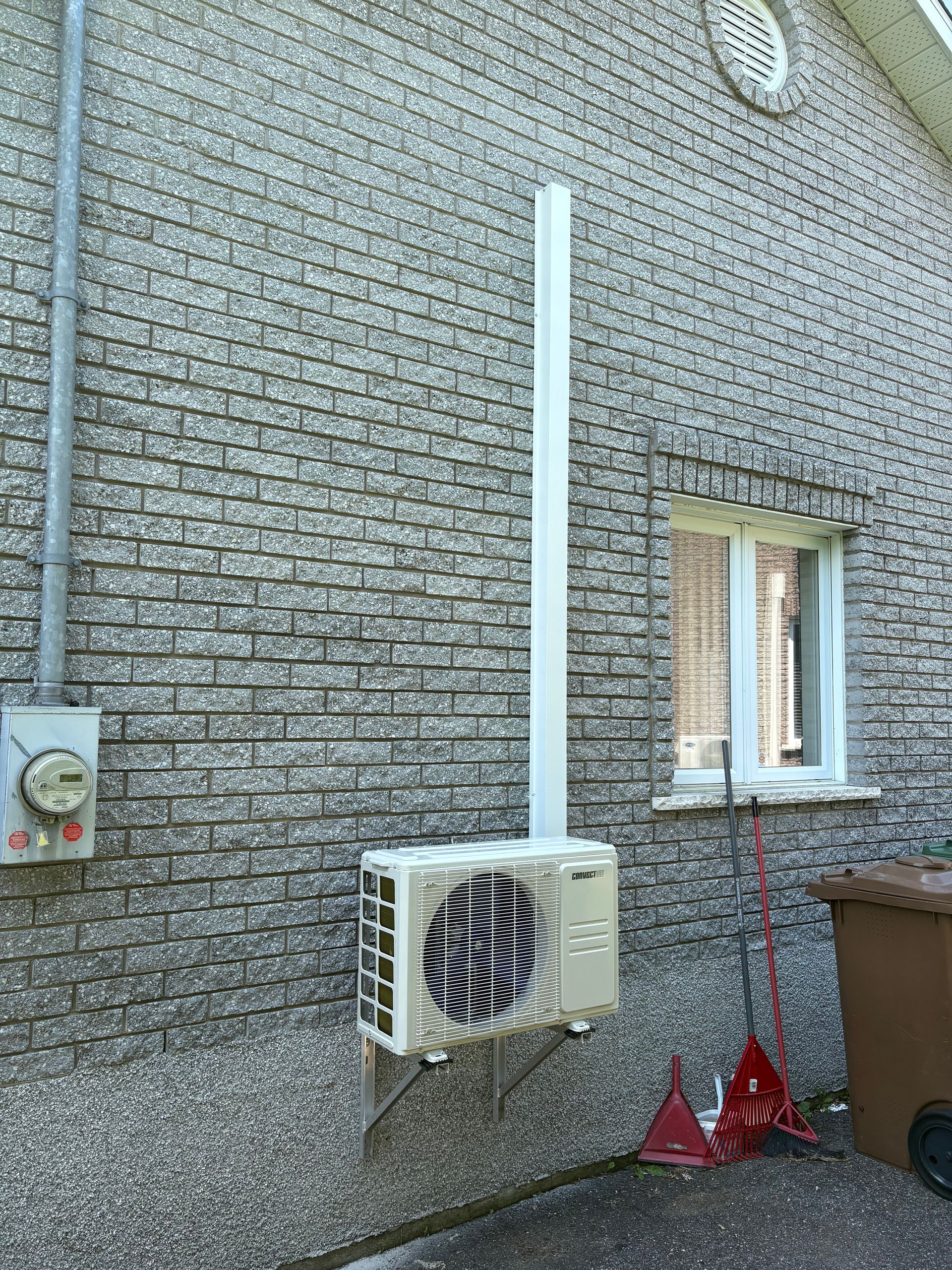 Un climatiseur extérieur fixé sur un mur de briques grises, avec un conduit blanc s'étendant vers le haut à côté d'une fenêtre.