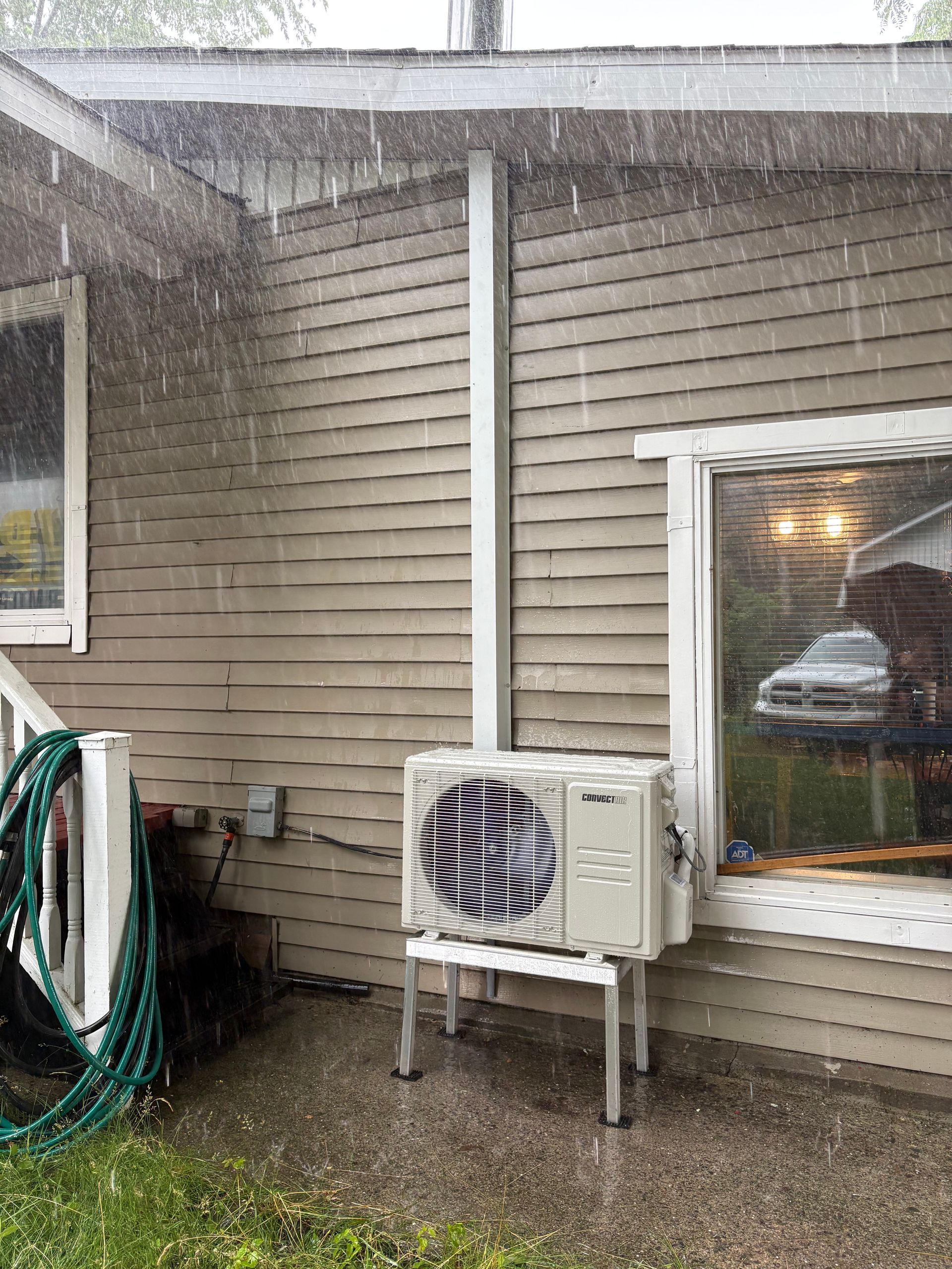 Une pompe à chaleur blanche se dresse sur un support métallique, adossée à un bardage beige, pendant un orage violent.