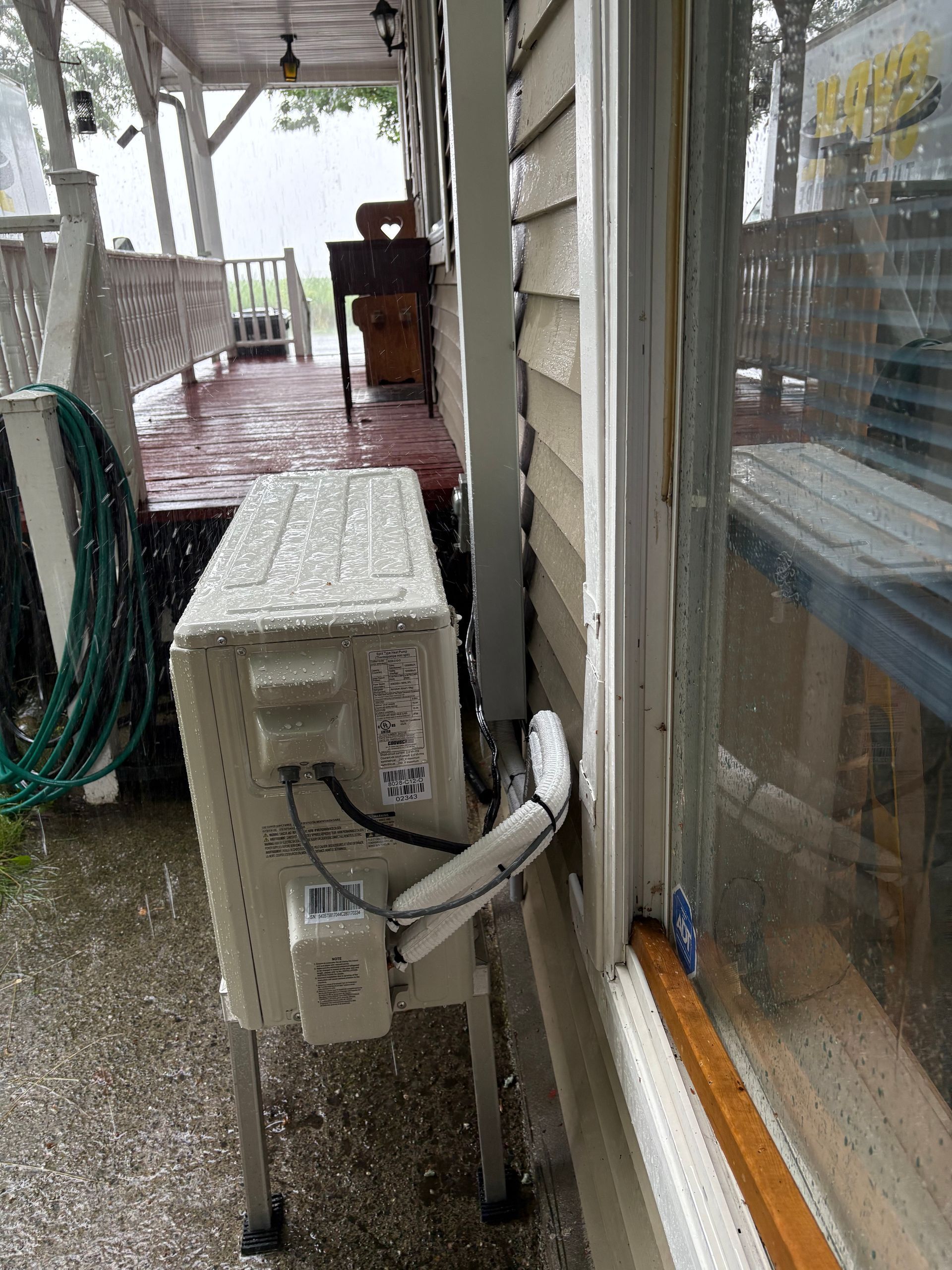 Une unité de climatisation extérieure blanche et rectangulaire repose sur des pieds à l'extérieur d'un bâtiment, partiellement obstruée par une fenêtre et un revêtement.