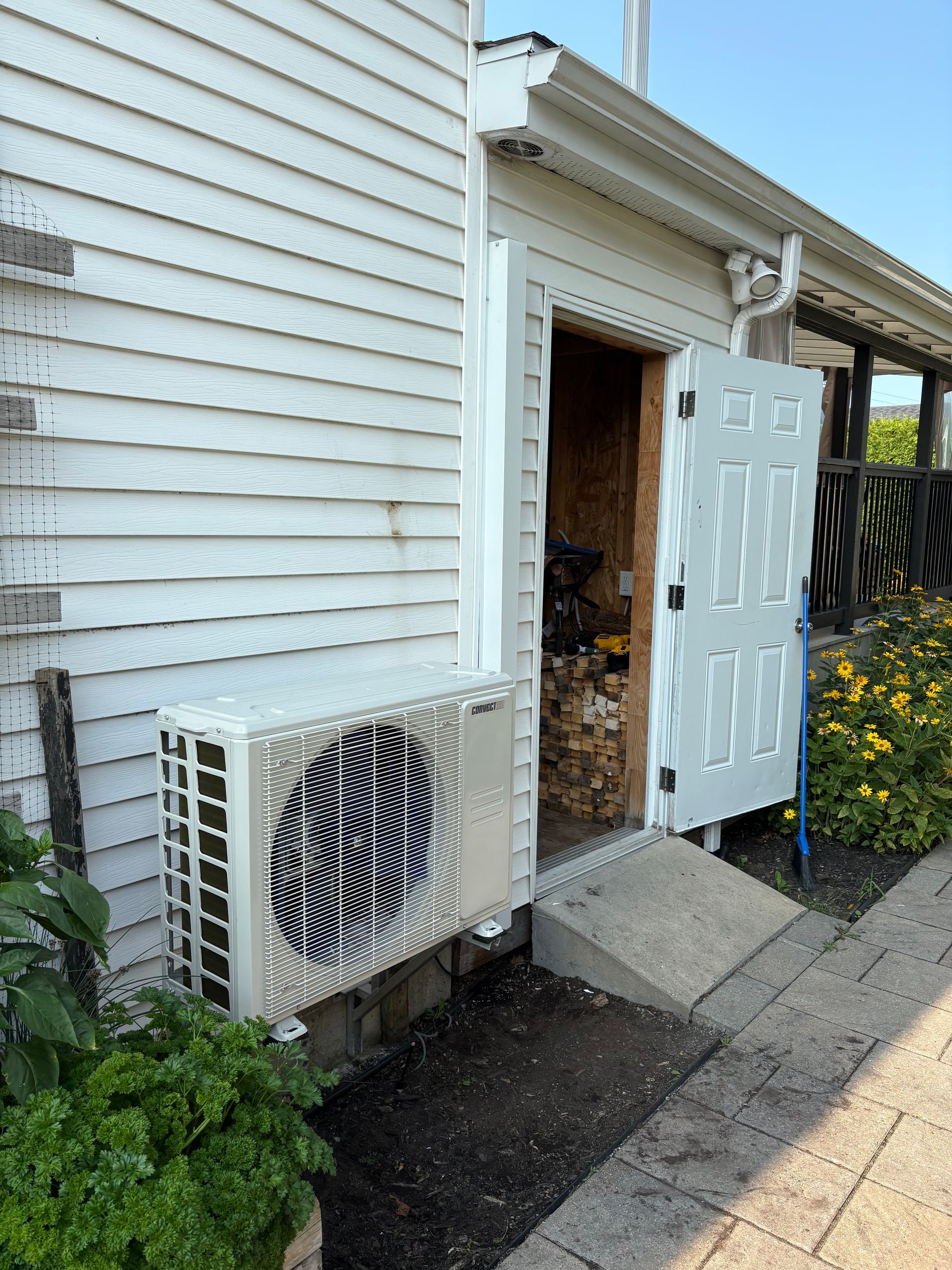 Un condenseur de climatisation blanc, monté sur le mur extérieur d'une maison, à côté de la porte ouverte d'un abri contenant du bois de chauffage.