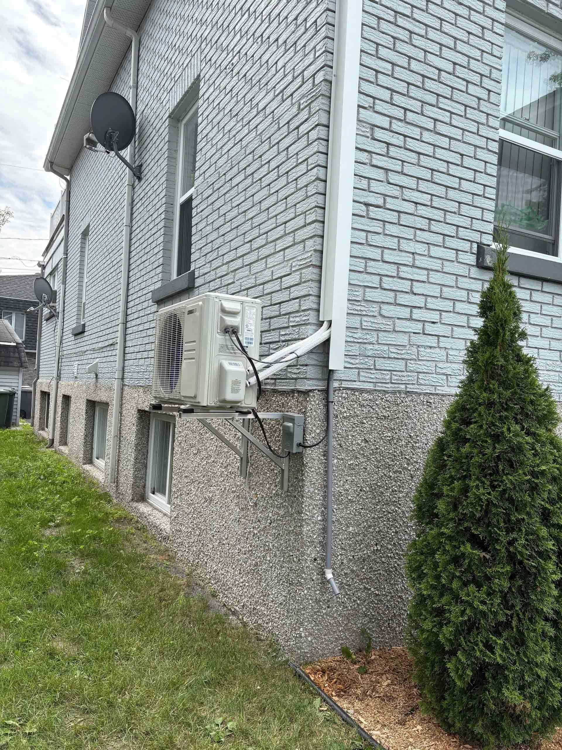 L'extérieur d'une maison en briques grises laisse apparaître un climatiseur mural et des fenêtres de sous-sol au-dessus de fondations en gravier.