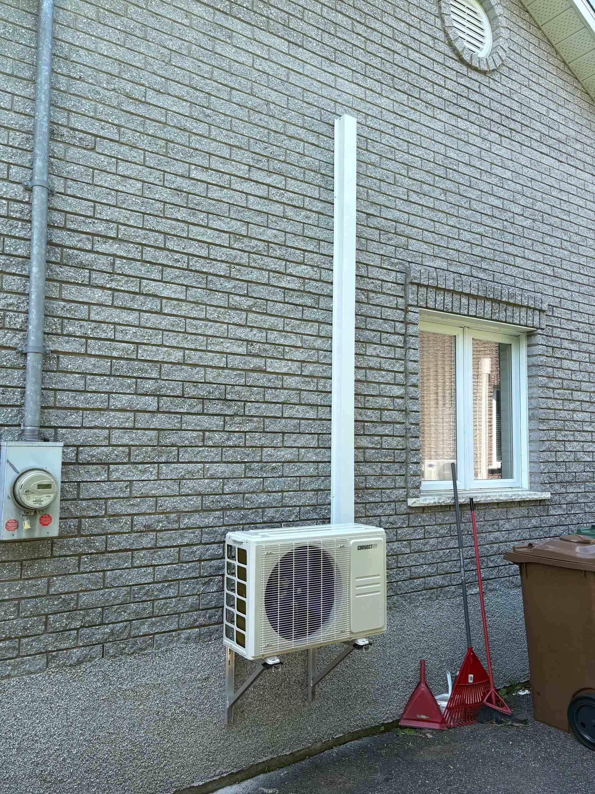 Un groupe de condensation de climatisation blanc, monté sur un mur extérieur en briques, avec un cache-conduit vertical blanc.