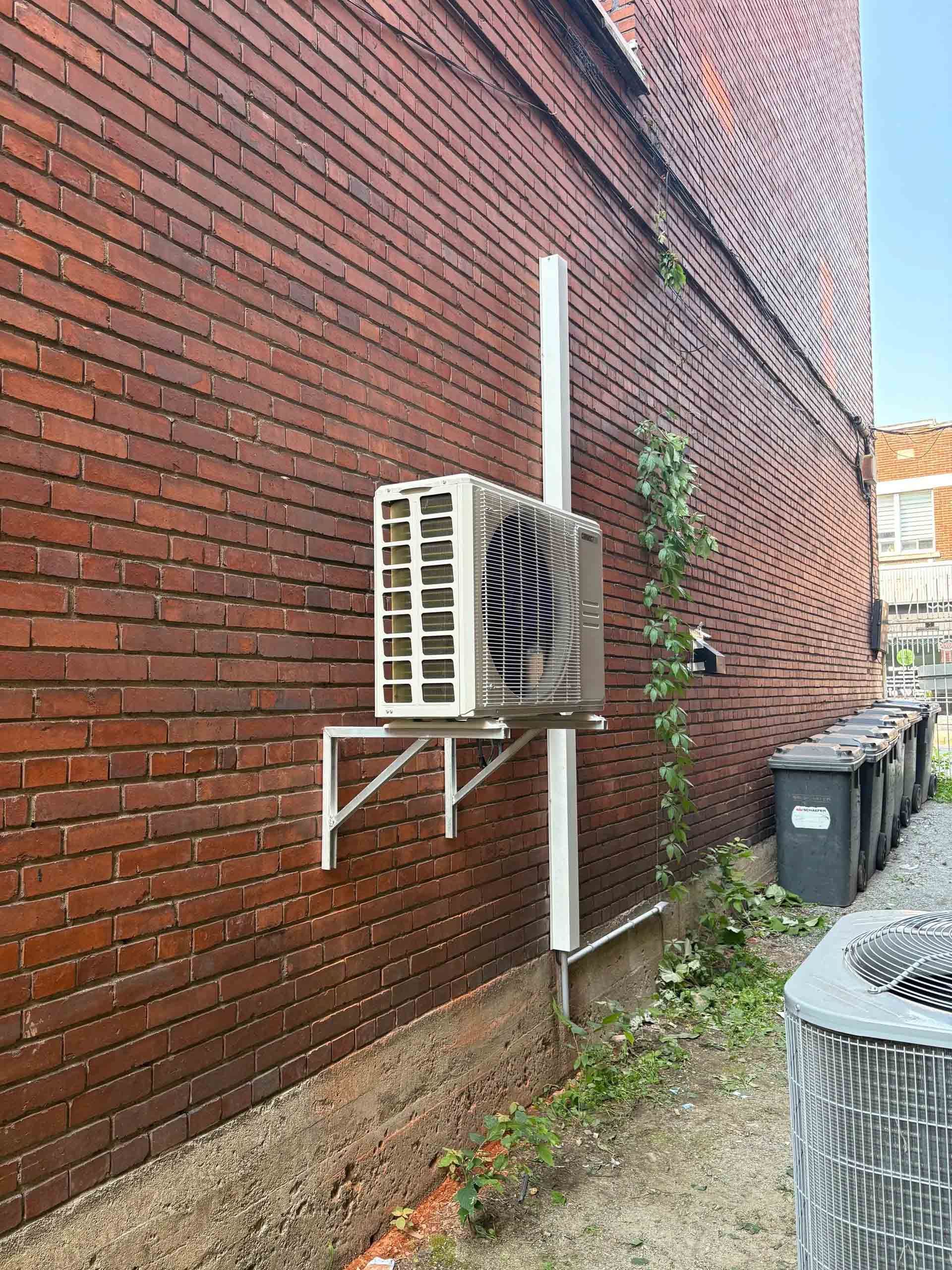 Un condenseur de climatisation blanc, monté sur des supports métalliques blancs contre un mur de briques rouges, avec un conduit vertical.