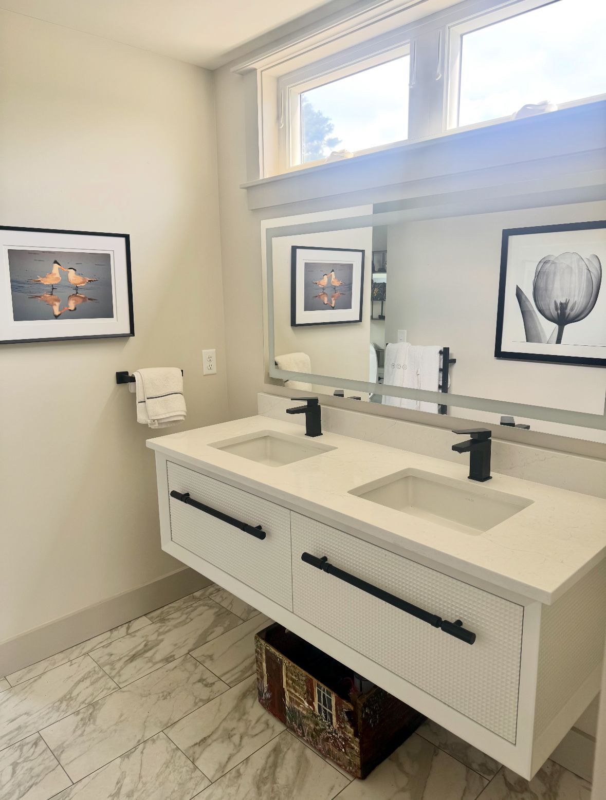 Bathroom with double sinks, gray walls, framed artwork, and a window. Black fixtures and a decorative basket.