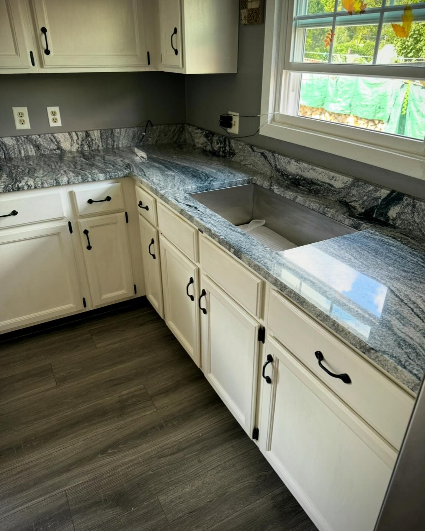 White kitchen cabinets with black hardware and a gray granite countertop, with a window view of greenery.