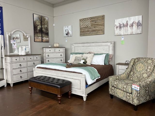 Bedroom furniture set with white bed, dresser, and patterned chair, dark wood floors.