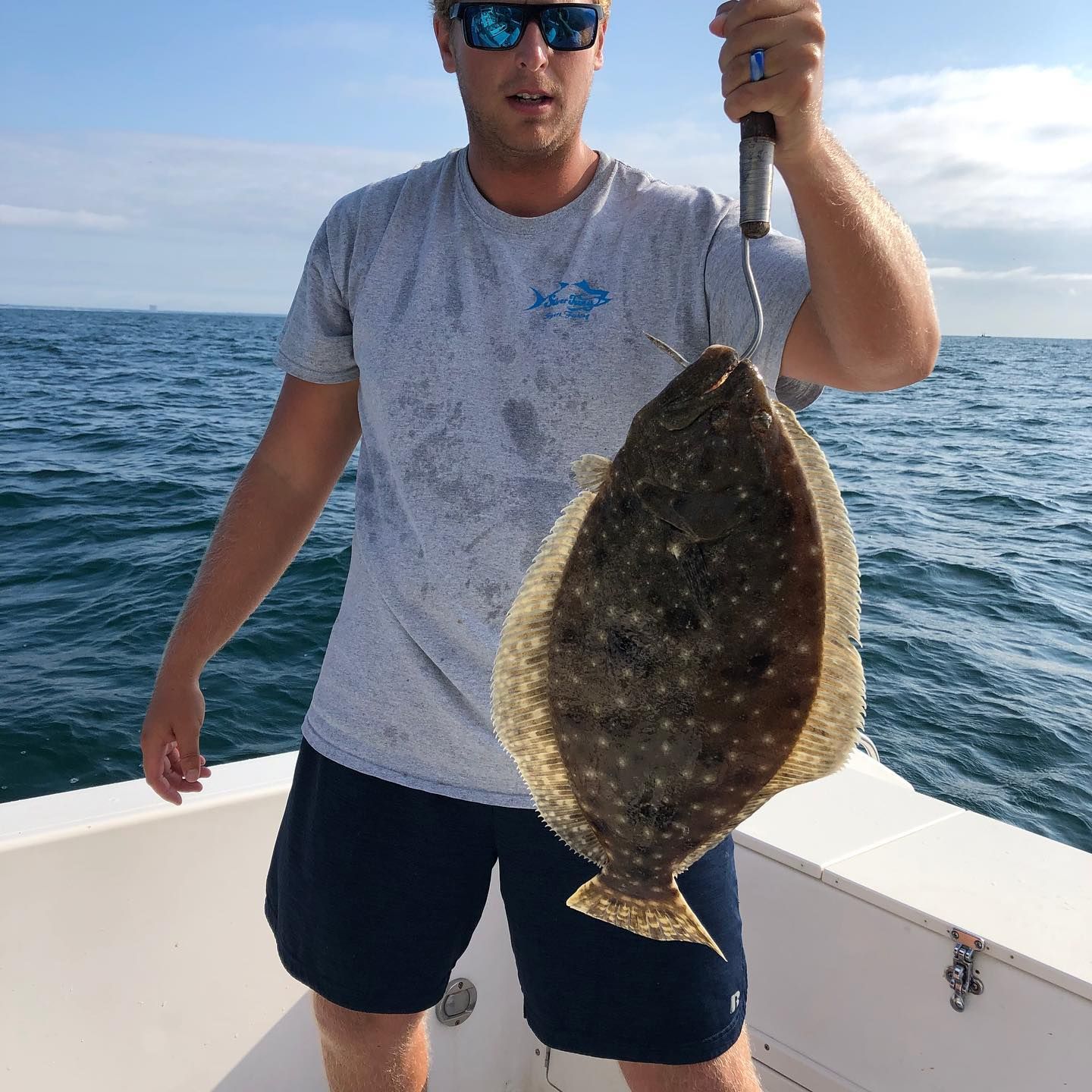 Man holding a flounder fish on a boat, blue sea in background. He wears sunglasses, a gray shirt, and navy shorts.