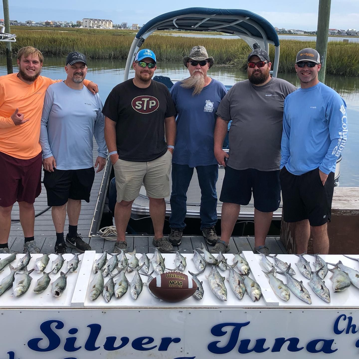 Seven men pose by a boat with fish on a table. The men are near water.