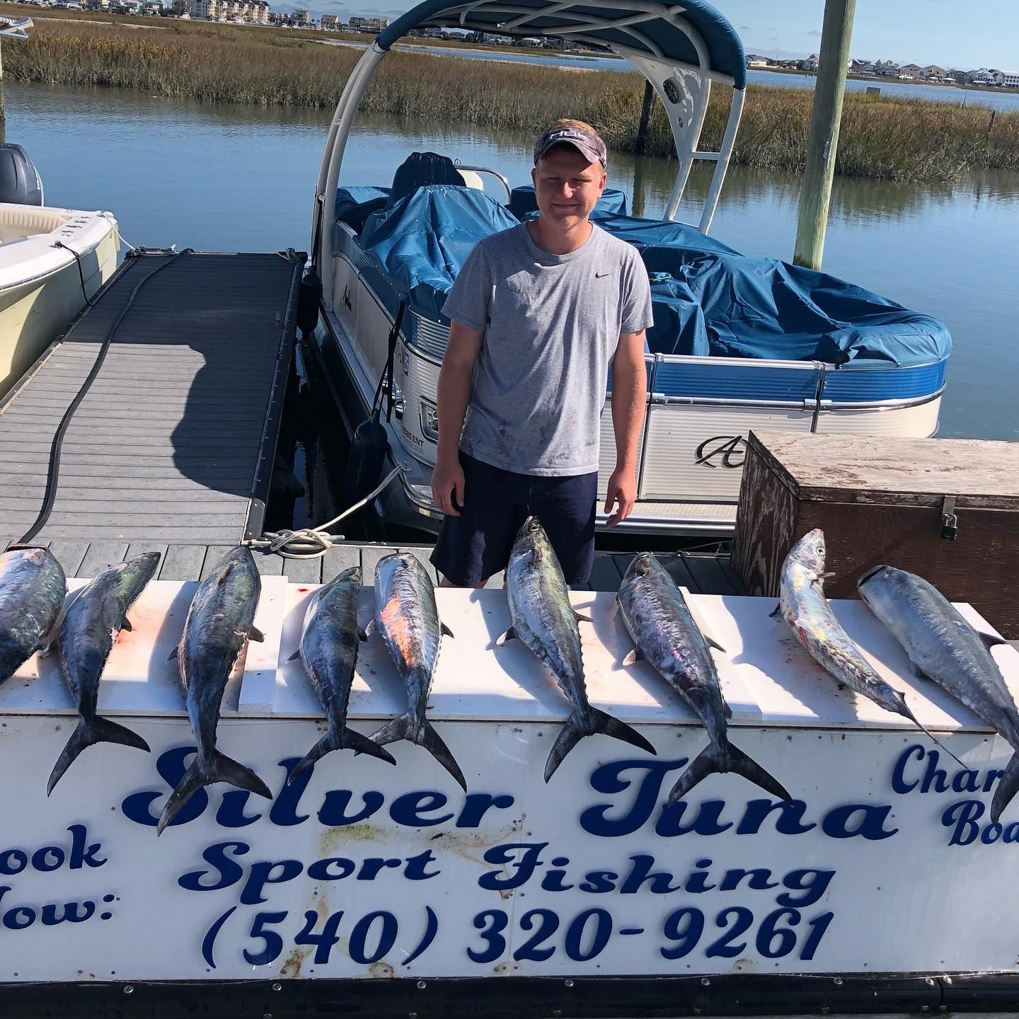 Man stands with caught silver tuna fish on dock, boat behind, river setting.