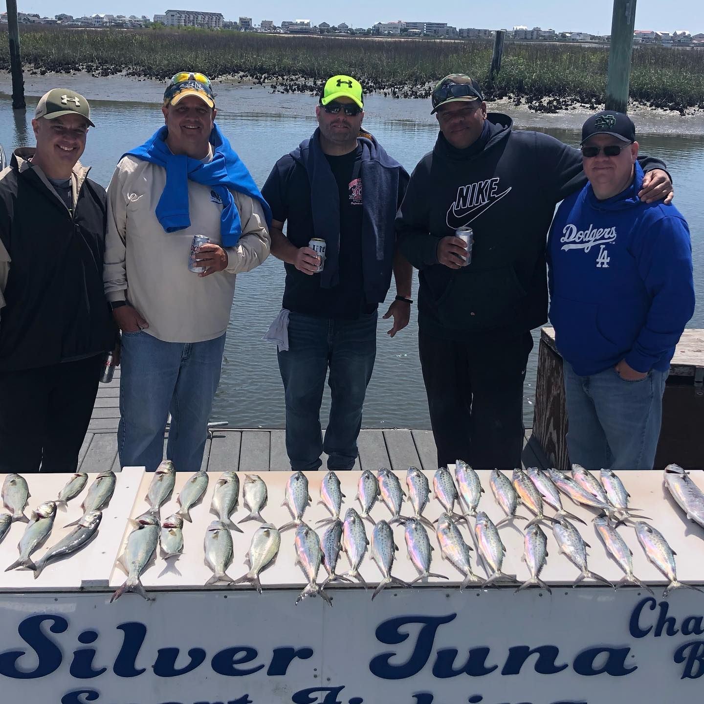 Five men stand on a dock with a large catch of fish, smiling. The dock is by water.