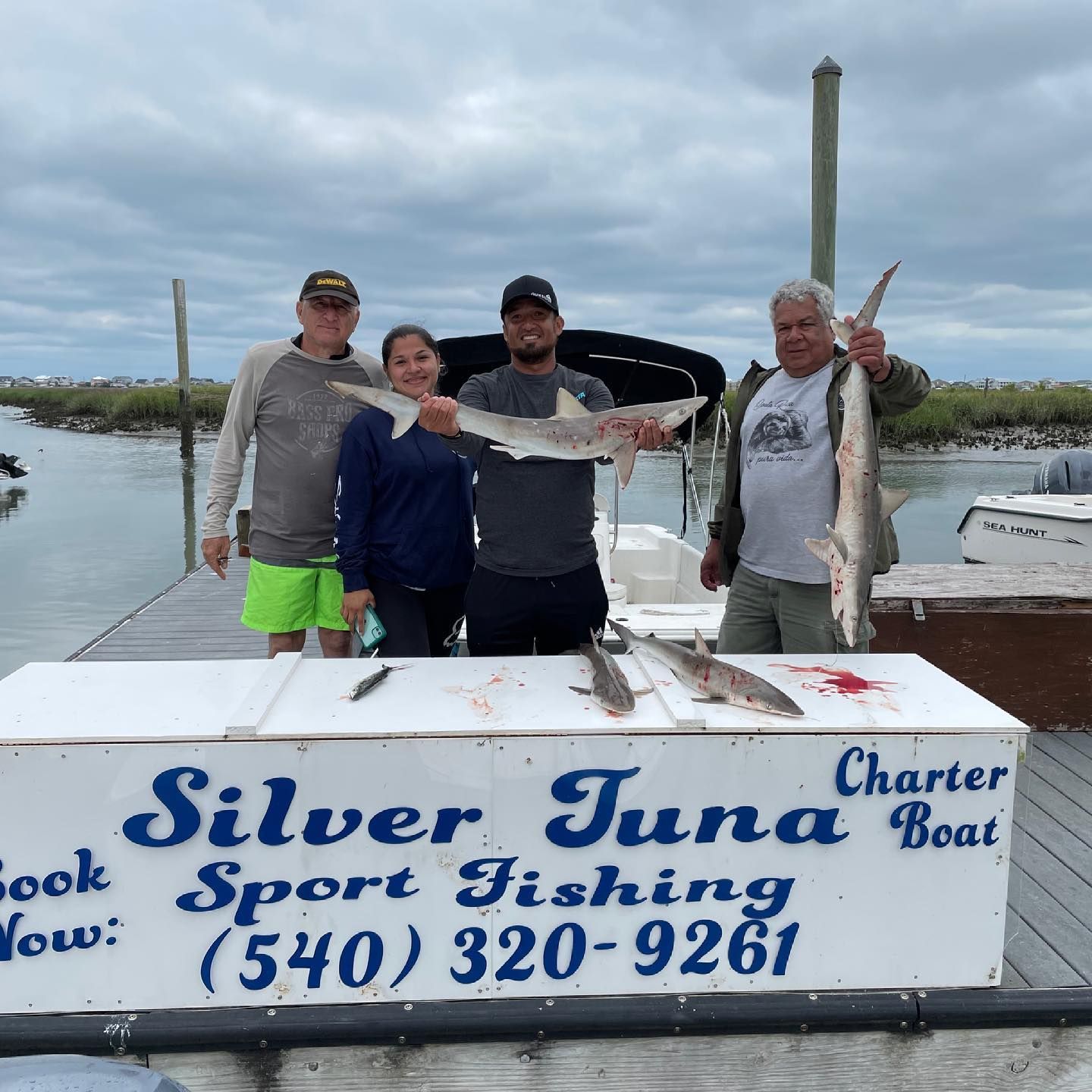 Four people on a dock with fish. The sign on the dock advertises 