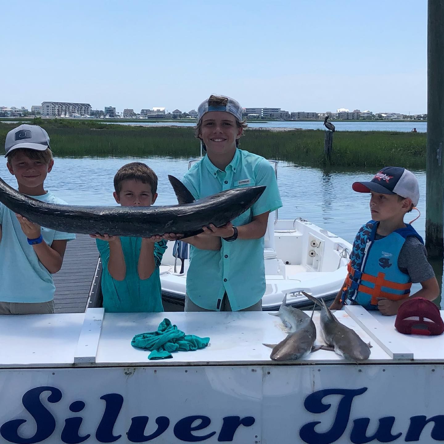 Four boys holding a large fish on a boat dock. Smiling, sunny day, buildings in background.
