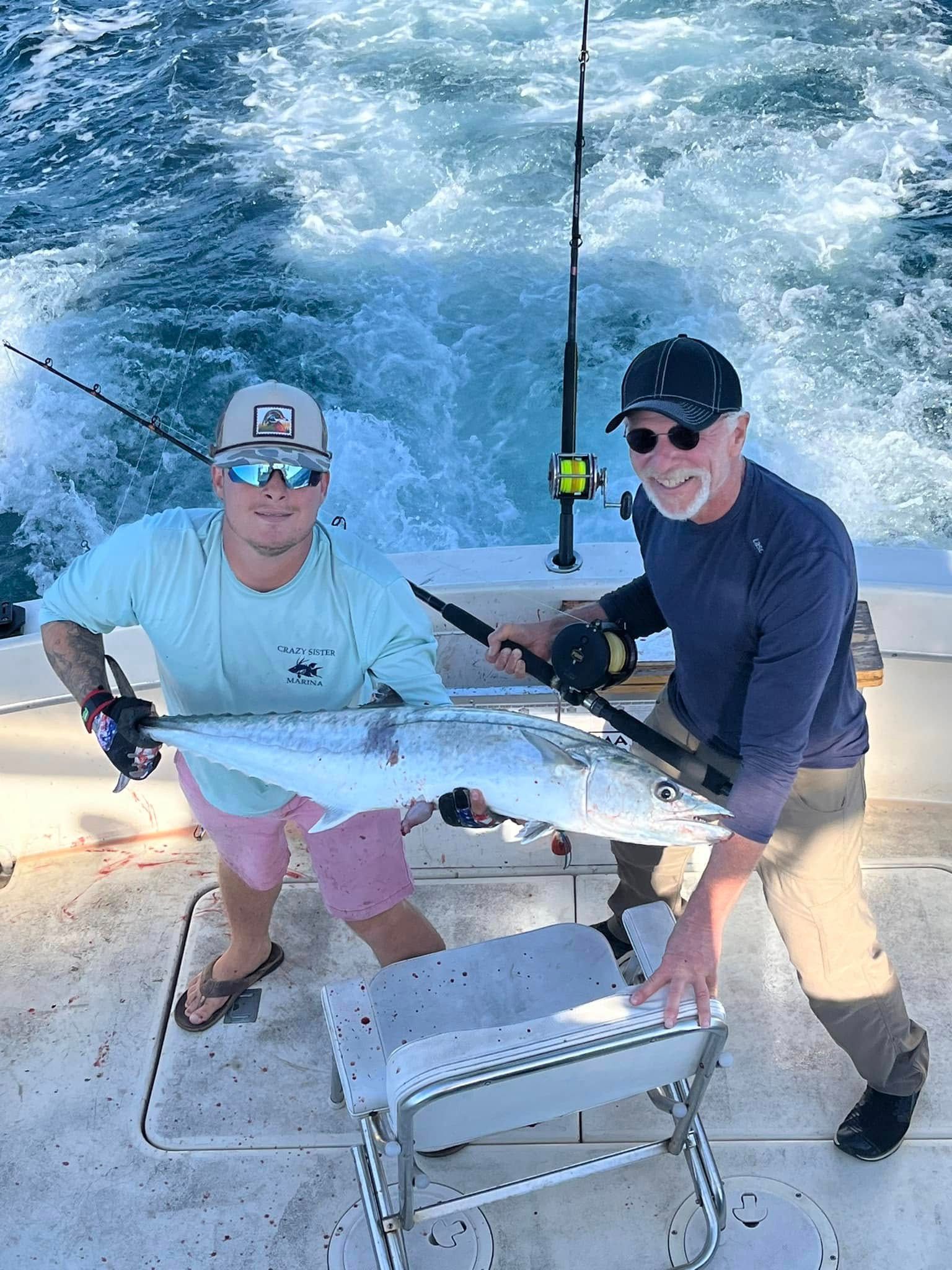 Two men on a boat holding a large silver fish; ocean waves behind them.