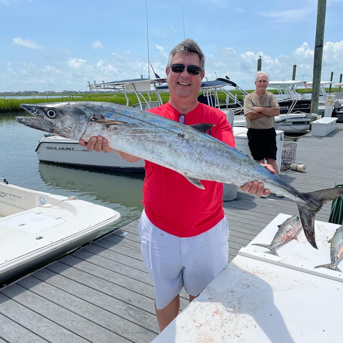 Man in red shirt holds a large fish on a dock near boats, another man in the background.