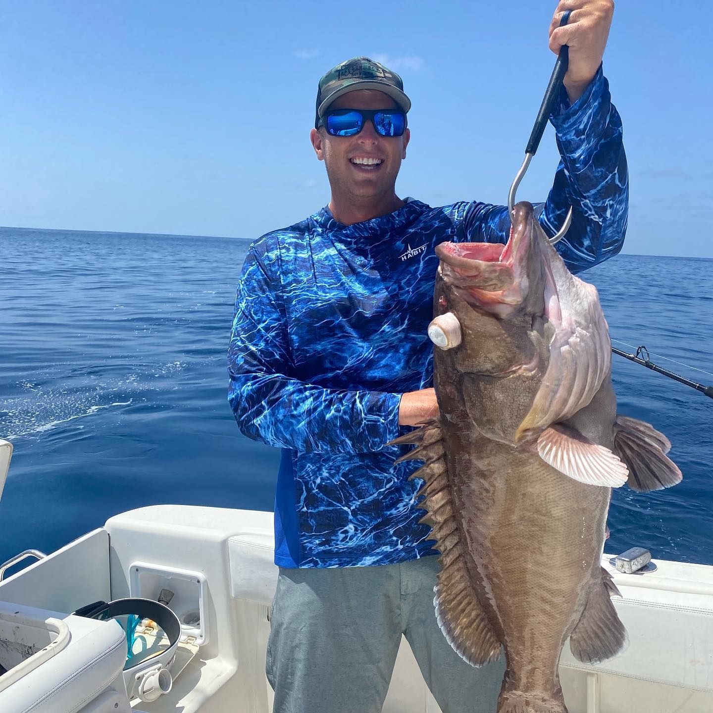 Man on boat holds up a large fish with mouth open, blue sea and sky.