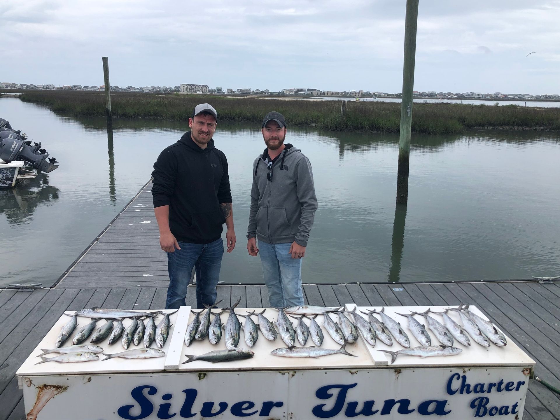 Two men on a dock with a display of caught fish. Cloudy day.