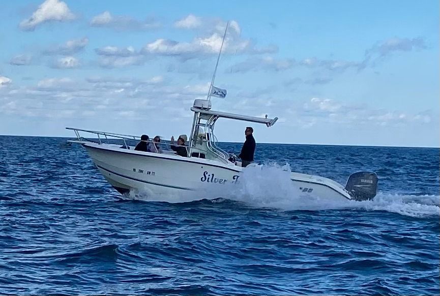 A white motorboat speeds across blue water with people on board under a partly cloudy sky.