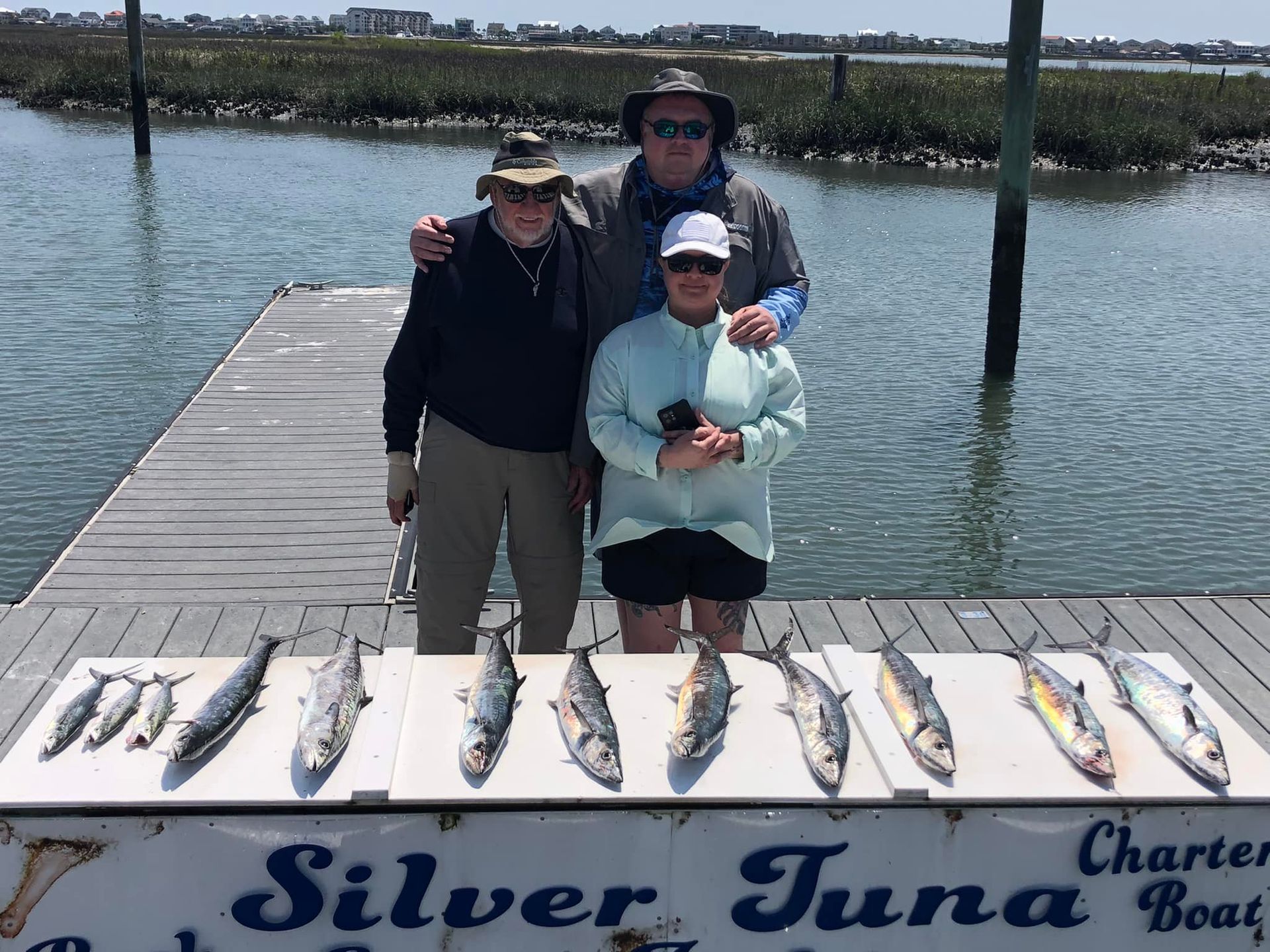 Three people pose with caught fish on a dock near the Silver Tuna charter boat.