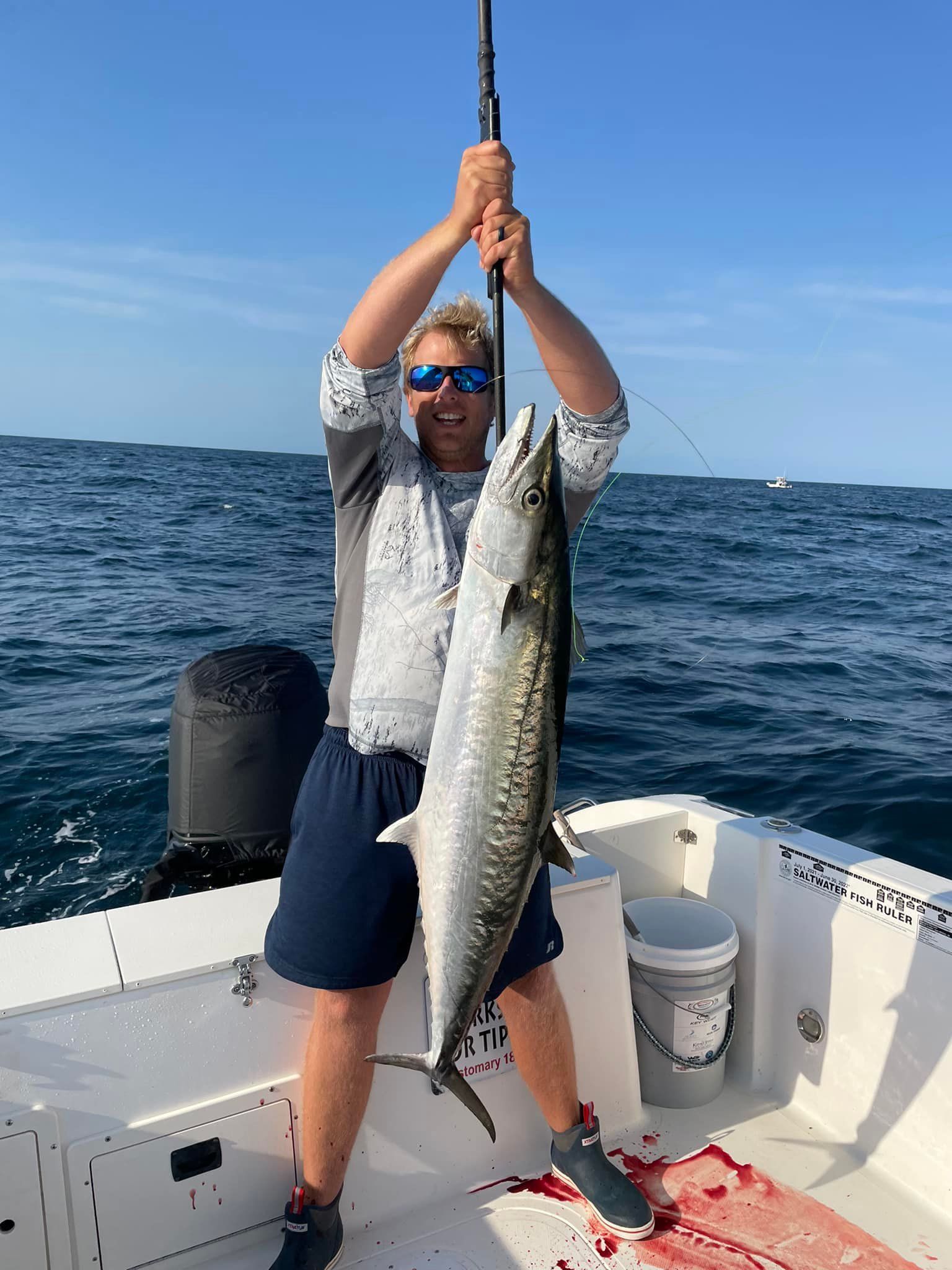 Man on a boat holds up a large fish, likely after catching it, under a blue sky.