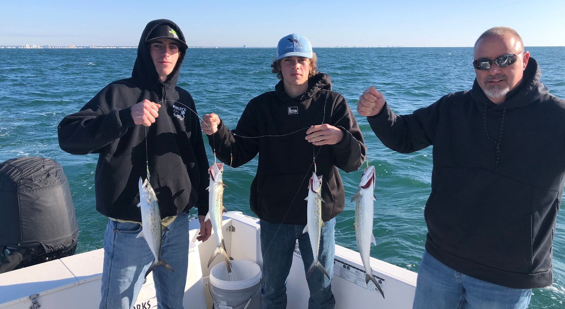Three people on a boat holding up fish they caught, smiling. Sunny day, water background.