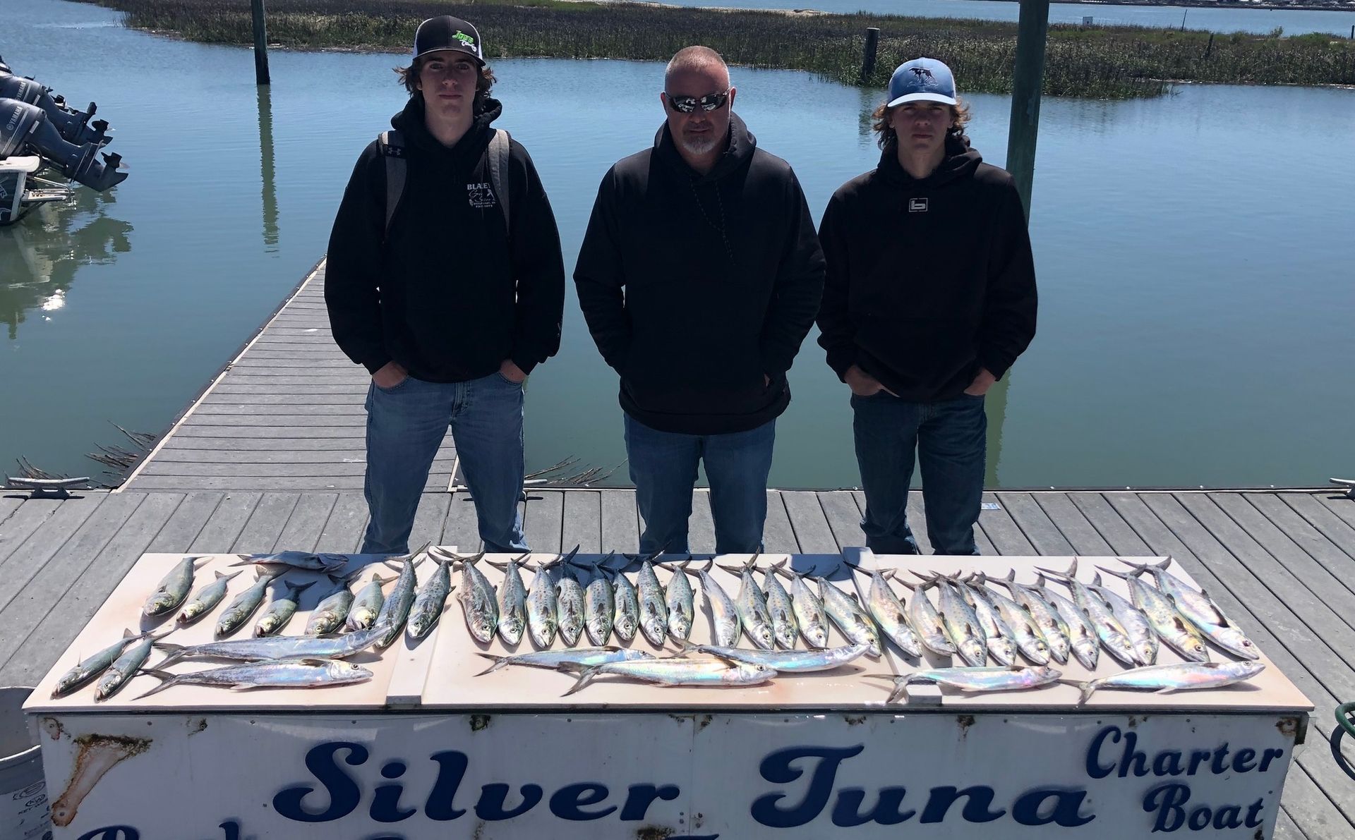 Three people pose on a dock with a table of fish; blue water and sky.