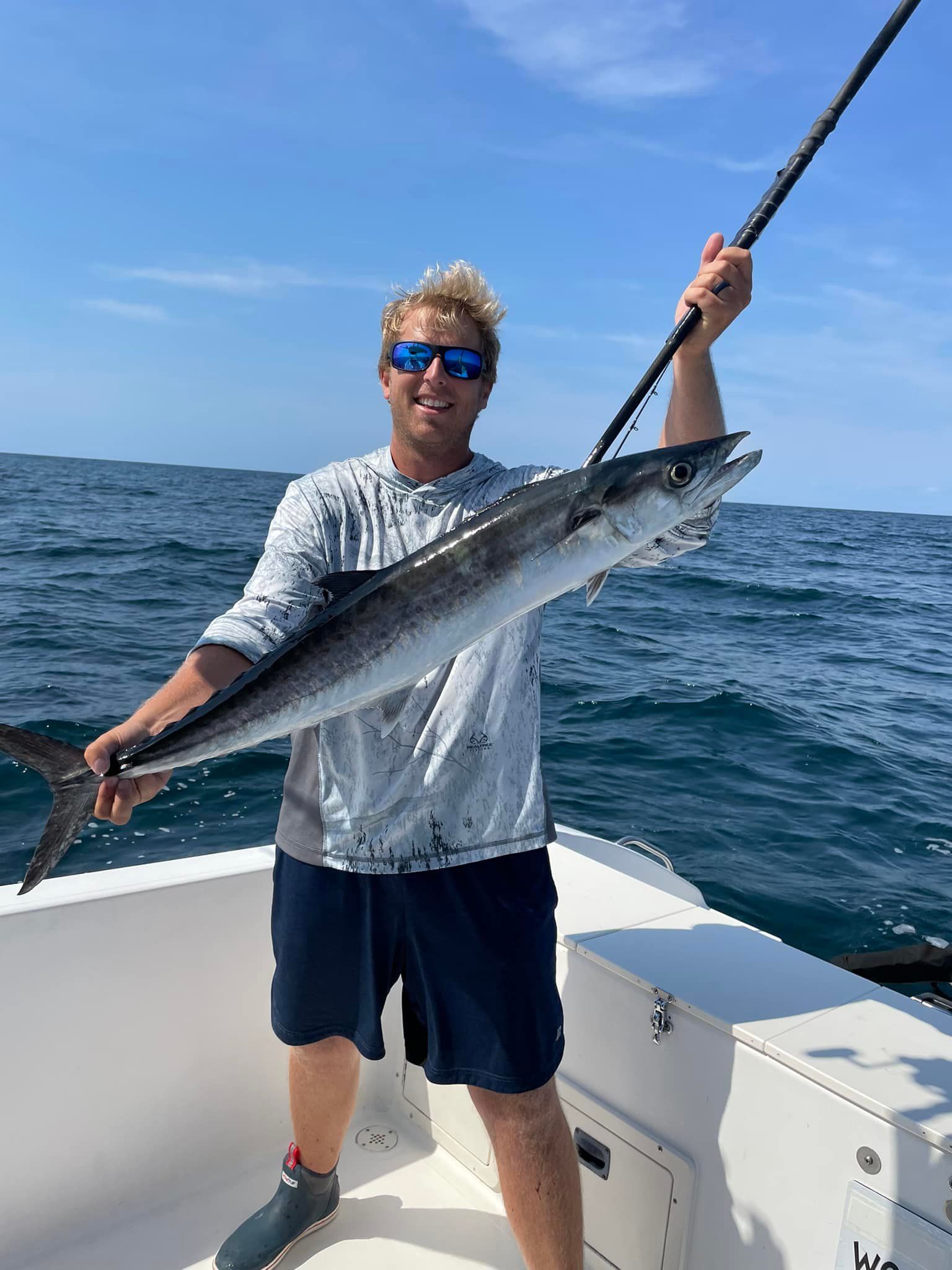 Man on a boat holds up a fish he caught, smiling. Blue sky and ocean in the background.