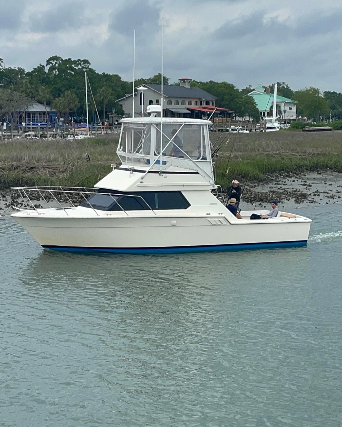 White boat with blue trim on water, fishing rods mounted, person at stern, background of buildings and trees.