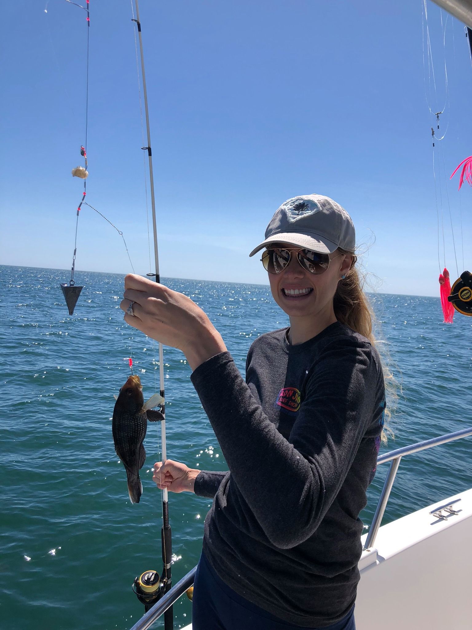 Woman fishing on a boat, holding up a small fish, smiling in the sun. Ocean and blue sky in background.