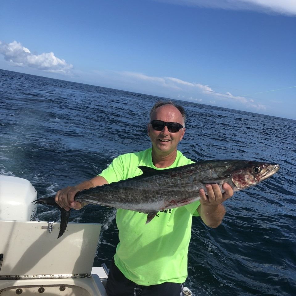 Man on a boat holding up a large king mackerel he caught. Blue water, sky; wearing a lime green shirt.
